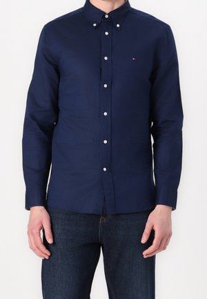 Mann trägt ein eng anliegendes, marineblaues, langärmeliges Hemd mit Knöpfen und einem kleinen Logo auf der Brust sowie dunkelblaue Jeans.