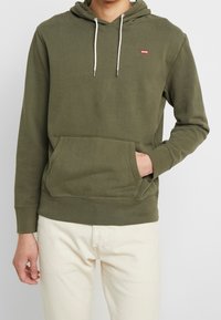 Sweat à capuche vert olive en mélange de coton avec une poche kangourou, une capuche à cordon de serrage, et un petit détail de logo rouge sur la poitrine. Finition en tissu texturé.