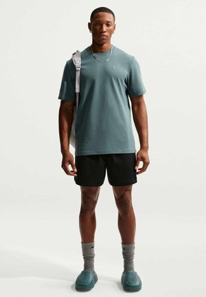 Mand iført grøn Nike-shirt, sorte shorts, grå sokker, grønne slip-on sko og bærer en rygsæk over den ene skulder, står stille.