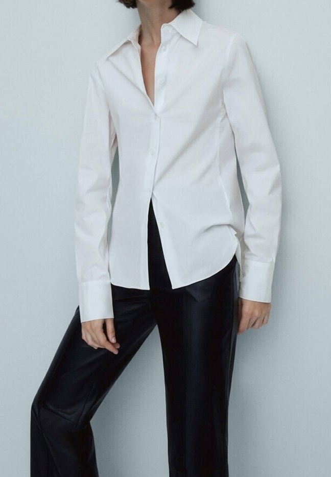 Femme portant une chemise blanche cintrée à boutons et un pantalon en cuir noir, debout devant un fond clair uni.