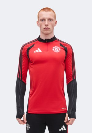 Mladý muž oblečený v červenom tréningovom tričku Manchester United s dlhým rukávom a čiernych Adidas nohaviciach, stojaci pred jednoliatym pozadím.