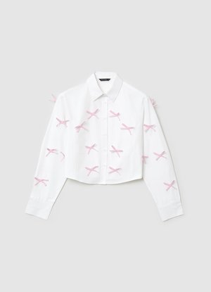Das weiße, kurze Hemd verfügt über lange Ärmel, eine Knopfleiste vorne und pinkfarbene Schleifenverzierungen in Kreuzmuster auf dem Stoff.