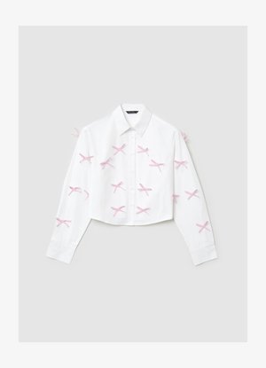 Das weiße, kurze Hemd verfügt über lange Ärmel, eine Knopfleiste vorne und pinkfarbene Schleifenverzierungen in Kreuzmuster auf dem Stoff.