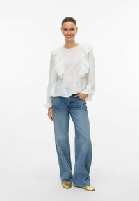 Blusa bianca con volant e maniche lunghe, vita arricciata e texture a righe sottili abbinata a jeans leggeri a vita alta di colore blu e ballerine dorate.