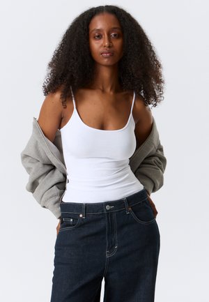 Femeie cu păr creț purtând un maiou alb, blugi închiși la culoare și o geacă gri, care alunecă de pe umeri, pe un fundal simplu și deschis la culoare.