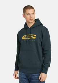 Ciemnozielona bluza z kapturem z sznurkiem i kieszenią z przodu, z żółtym logo G-Star i napisem. Wykonana z miękkiej mieszanki bawełny, ma casualowy design.