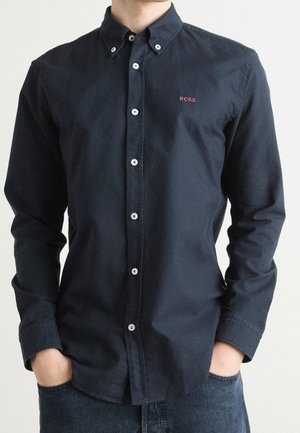 Mann trägt ein dunkelblaues Hemd mit Knöpfen, auf der linken Brust rot mit "BOSS" bestickt, Hände in den Jeans-Taschen.