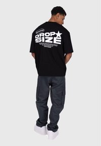 Svart oversized t-skjorte med hvit grafisk tekst på baksiden, kombinert med grå cargo-bukser og hvite sneakers. Minimalistisk design, avslappet passform.