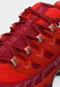 Κόκκινο αθλητικό παπούτσι με υφή στην επάνω επιφάνεια, περίπλοκο σύστημα κορδονιών και αντιθετικές λεπτομέρειες. Αναδεικνύεται από γεωμετρικά σχέδια και ανθεκτική σόλα.