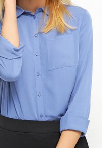 Camisa de botones de manga larga azul claro con un bolsillo en el pecho y los puños doblados, hecha de una tela suave, combinada con una falda negra.
