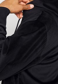 Svart sammetjacka med en slät textur, ribbade muddar och dragsko i huvan, som visar detaljer av tyget och en hand som justerar kragen.