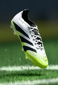 Crépins de football blancs avec des rayures noires, une tige texturée, des accents jaunes sur la semelle et un col en flyknit, présentés sur de l'herbe avec des débris volant.