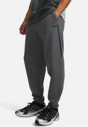 Man draagt losse grijze Under Armour joggingbroek en zwarte sneakers, staat met één hand in de zak, gedeeltelijk zicht op bovenlichaam en benen.