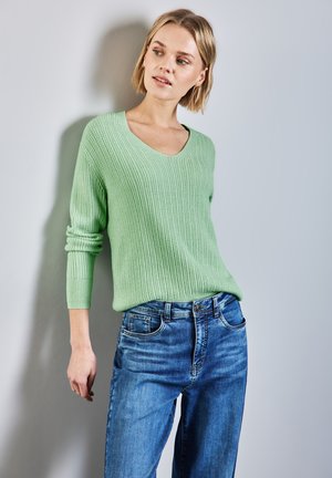 Hellgrüner gerippter Pullover mit V-Ausschnitt und langen Ärmeln, kombiniert mit hoch taillierten blauen Jeans, die eine lockere Passform und eine weiche Textur aufweisen.