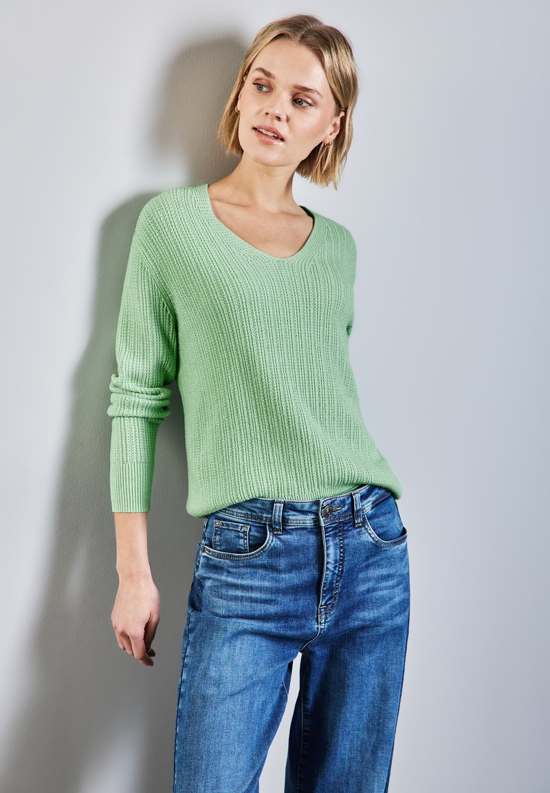 Maglione a coste verde chiaro con scollo a V e maniche lunghe, abbinato a jeans blu a vita alta, che mostrano una vestibilità comoda e una consistenza morbida.