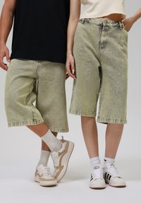 Light green denim knee-length shorts with a faded texture, featuring prominent stitching and a relaxed fit. Paired with white sneakers.