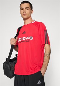 Punainen urheilupaita mustalla Adidas-logolla, lyhyet hihat, pyöreä kaula-aukko ja mustat raidat hihoissa. Yhdistetty mustiin shortseihin ja laukkuun.
