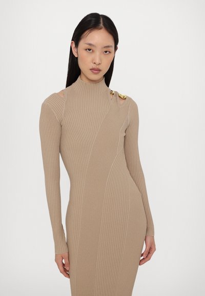 Knit dress in light beige with ribbed texture, high neck, long sleeves, and decorative gold hardware on the shoulder. Form-fitting design.