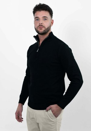Zwarte zip-up trui met een hoge kraag, gemaakt van een zachte stof. Heeft een slim fit en geribde mouwen, gecombineerd met lichtgekleurde broeken.