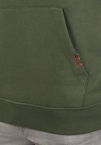 Maslinasto zeleni dukserica s kengurua džepom, rebrastim donjim rubom i malim smešnim kožnim etiketom s logotipom sa strane. Mekani pamučni materijal.