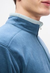 Ανοιχτό μπλε fleece μπουφάν με ψηλό γιακά και αντίθετο εσωτερικό γιακά σε ανοιχτό μέντα πράσινο. Διαθέτει φερμουάρ και ραμμένες λεπτομέρειες.