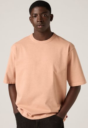 Jeune homme à la peau foncée et aux cheveux courts portant un t-shirt ample couleur pêche clair et un pantalon noir, les mains dans les poches.