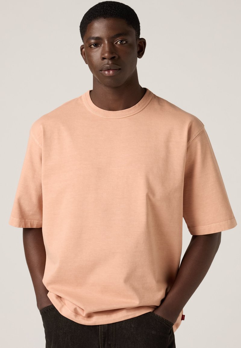 Junger Mann mit dunkler Haut und kurzen Haaren, der ein helles pfirsichfarbenes oversized T-Shirt und schwarze Hosen trägt, die Hände in den Taschen.