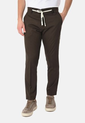 Uomo che indossa pantaloni slim fit marrone scuro con cintura bianca a cordoncino e sneakers in suede taupe, in piedi con le mani nelle tasche.
