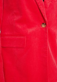 Raudonas kordūrojo švarkas su vieno mygtuko uždarymu, turintis vertikalių raukšlių tekstūrą ir šoninį kišenę.