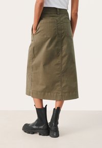 Olivgrünen Baumwoll-Midi-Rock mit zwei Gesäßtaschen, flachem Bund und seitlichen Schlitzen. Kombiniert mit schwarzen Ankle Boots mit dicken Sohlen.