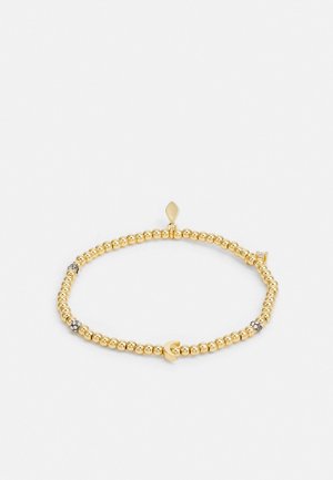 Pulsera de cuentas doradas con pequeñas cuentas con acentos plateados y un colgante en forma de corazón sobre un fondo blanco liso.