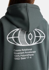 Dunkelgrüne Kapuzenjacke mit einem großen weißen Grafikdesign auf der Rückseite, Text, der Material und Modell beschreibt, einfarbig und mit weicher Textur.