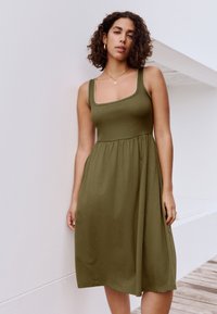 Olivengrøn kjole med tætsiddende krop, firkantet halsudskæring og udvidet skørt. Ribbet tekstur med dekorativ kant langs halsudskæringen.