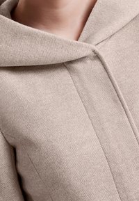 Beige huva jacka tillverkad av texturerad tyg, med en strömlinjeformad design, synliga sömmar och dragkedja fram.