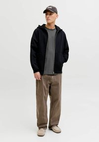 Sweat à capuche noir zippé avec deux poches avant, porté par-dessus un t-shirt gris, associé à un pantalon marron ample et des chaussures beige sans lacets. Casquette sombre sur la tête.