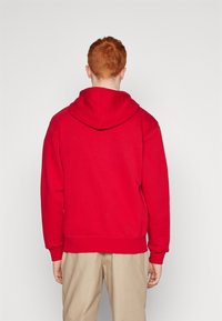 Jongere met rood haar die een eenvoudige rode hoodie en beige broek draagt, gezien van de achterkant tegen een witte achtergrond.