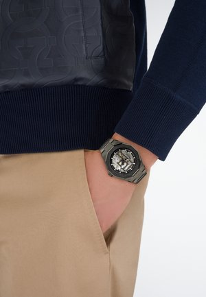 Personne portant un pull bleu foncé avec la main dans la poche d’un pantalon beige, montrant une montre-bracelet en métal noir avec un mécanisme d’engrenage visible.