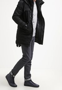 Svart puffjacka med luva, grå tröja och grå jeans. Mörkblå högstövlade sneakers med texturerad yta och kontrasterande detaljer.