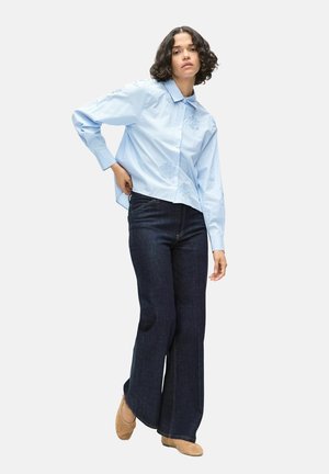 Femme aux cheveux courts et bouclés portant une chemise bleu clair boutonnée, un jean évasé foncé et des chaussures beiges, debout devant un fond uni.