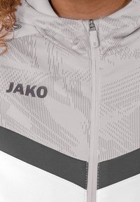 Giacca zip-up bianca e grigia con motivo geometrico, logo JAKO e strisce grigie a contrasto lungo la zona del petto. Tessuto dalla texture liscia.