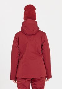 Röd isolerad jacka med huva, med en slät yta, sömmar som löper ner längs ryggen och en matchande röd stickad mössa.
