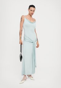 Een lichtblauwe, mouwloze maxi-jurk met een gedrapeerde halslijn, vastgebonden in de taille. Gecombineerd met een zwarte handtas en crèmekleurige puntige hakken.