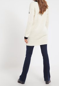 Sweter w kolorze kremowym z dzianiny, z prążkowanym dołem i czarnymi akcentami, w połączeniu z ciemnoniebieskimi spódnicami dzwonowymi, widoczny od tyłu.