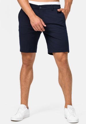 Mand iført sorte skræddersyede shorts og hvide sneakers står med hænderne nær lommerne på en ensfarvet baggrund.
