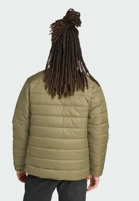 Person med lange dreadlocks, der vender væk, iført en olivengrøn pufferjakke, mod en lys grå baggrund.