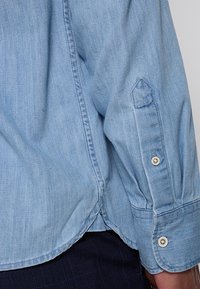 Camicia in denim azzurro chiaro con polsini abbottonati, colletto a punta e orlo curvo con dettagli di cucitura. Texture liscia e design informale.