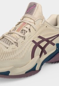 Κοντινό πλάνο μπεζ αθλητικού παπουτσιού Asics με μοβ λογότυπο, υφασμάτινο πάνω μέρος με υφή, κρεμ κορδόνια και μπλε και μοβ λεπτομέρειες στη σόλα.