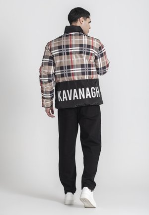 Mann trägt eine beige-schwarz karierte Jacke mit dem Text "KAVANAGH" auf dem Rücken, schwarze Hose und weiße Sneaker, steht mit dem Rücken zur Kamera.