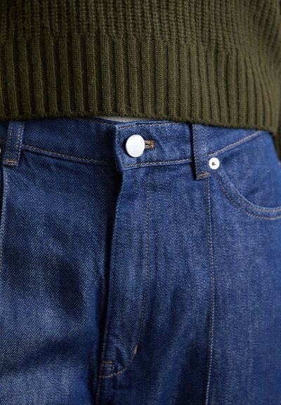 Close-up of blue denim jeans buttoned at the waist beneath an olive-green ribbed sweater.