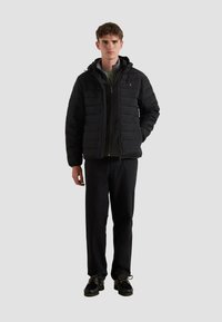 Giacca nera imbottita con zip e cappuccio, caratterizzata da un design a righe verticali. Indossata sopra una maglia scura e abbinata a pantaloni e scarpe scure.